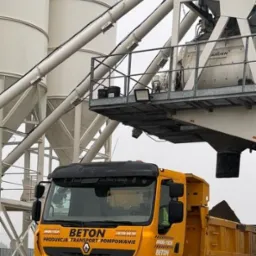Żółta betonomieszarka Renault z napisem 'BETON' na tle silosów i konstrukcji betoniarni.