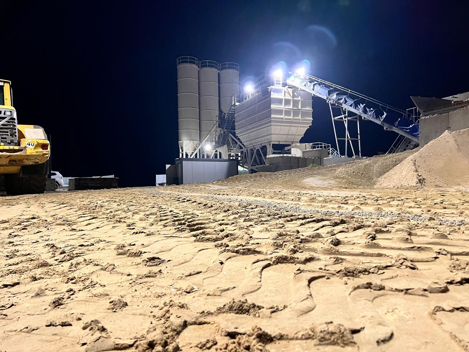Oświetlona nocą instalacja do produkcji mieszanek betonowych z widocznym podajnikiem taśmowym i stertą kruszywa, z żółtą ładowarką kołową w tle po lewej stronie, na pierwszym planie ziemia...