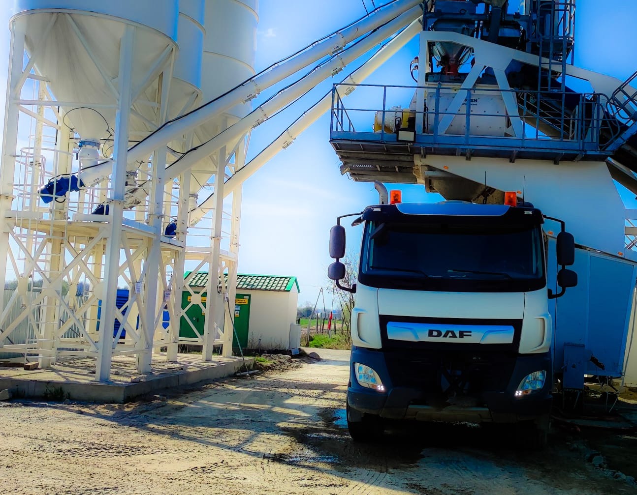 Ciężarówka DAF na tle białej konstrukcji betoniarni z silosami i zielonego budynku gospodarczego w słoneczny dzień.