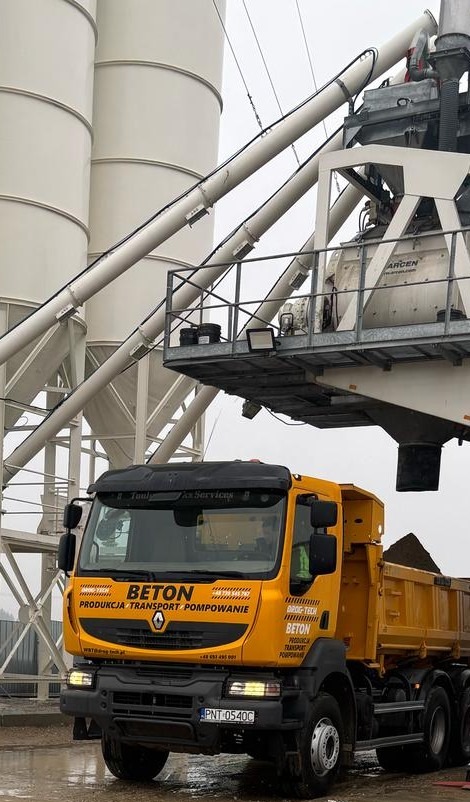 Żółta betonomieszarka Renault z napisem 'BETON' na tle silosów i konstrukcji betoniarni.