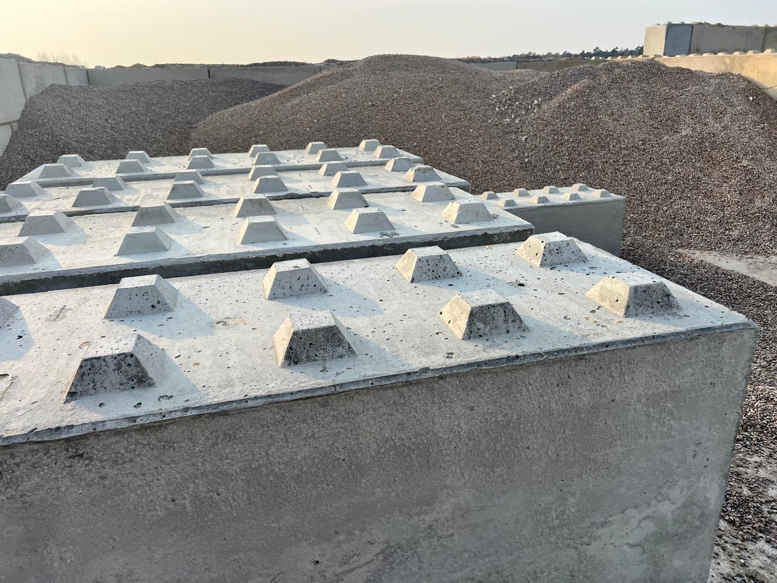 Szare betonowe bloki z piramidowymi wypustkami na górze, ułożone w rzędach, w tle hałda żwiru i inne bloki.