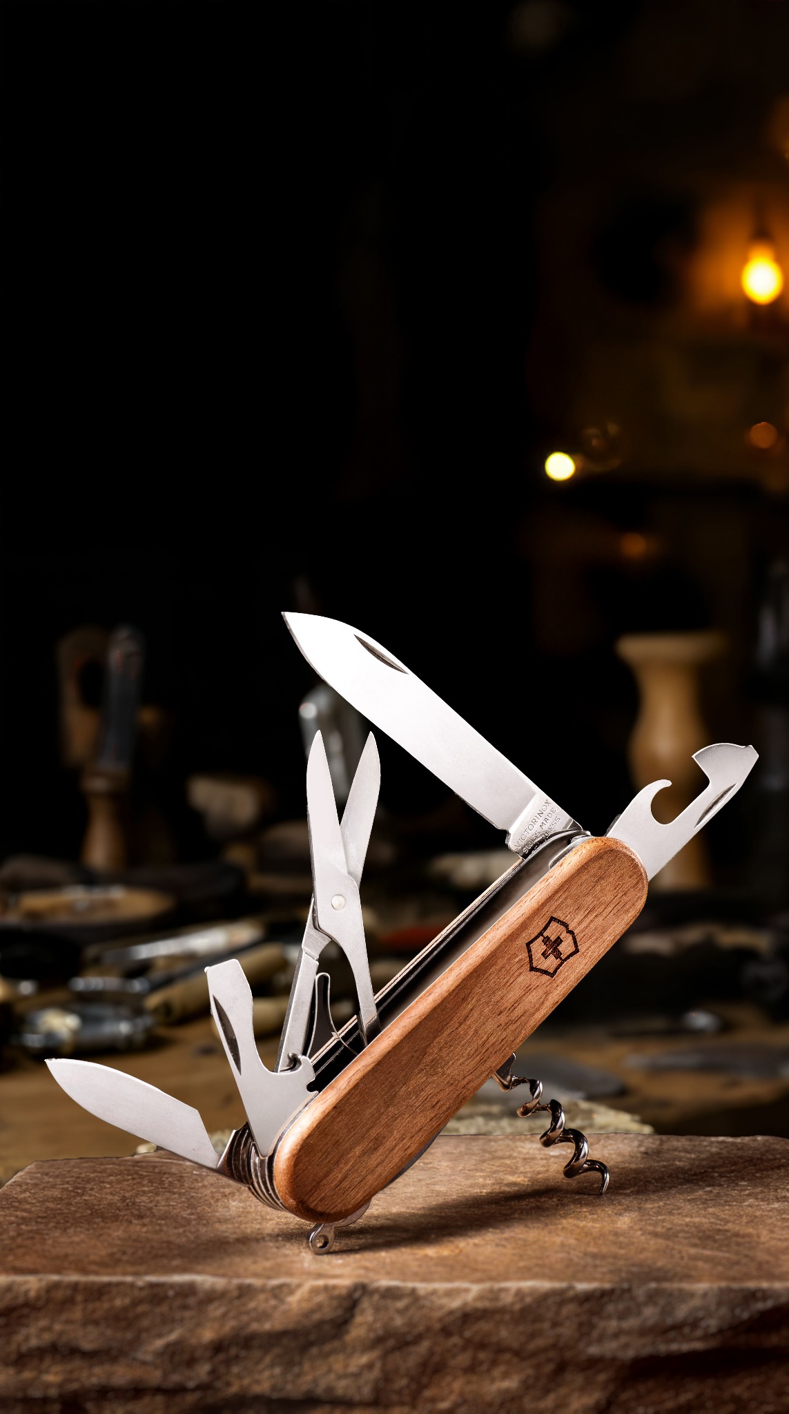 Otwarty scyzoryk szwajcarski z drewnianą rękojeścią, ukazujący ostrze, nożyczki i korkociąg, umieszczony na kamiennej powierzchni na rozmytym tle z narzędziami i oświetleniem.