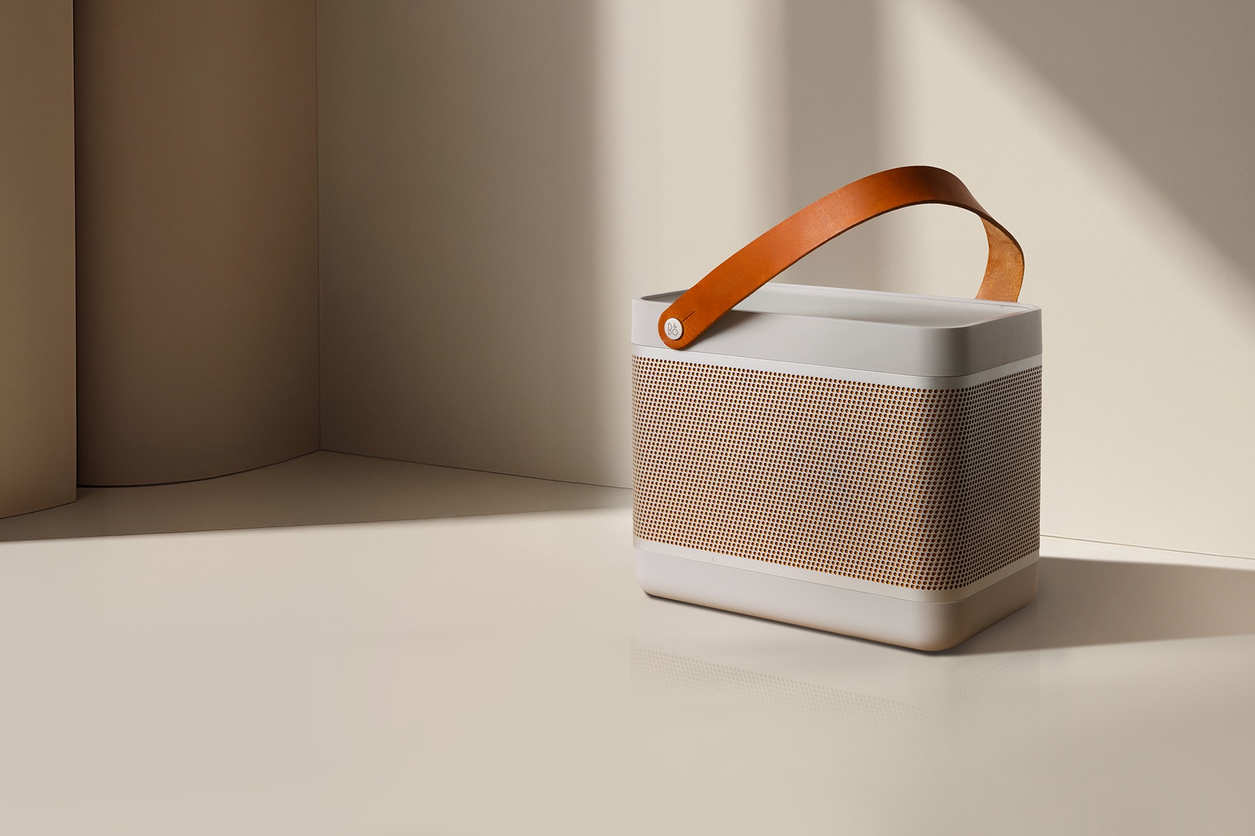 Szary głośnik przenośny z perforowaną złotą siatką i skórzanym uchwytem na beżowym tle z delikatnym cieniem.