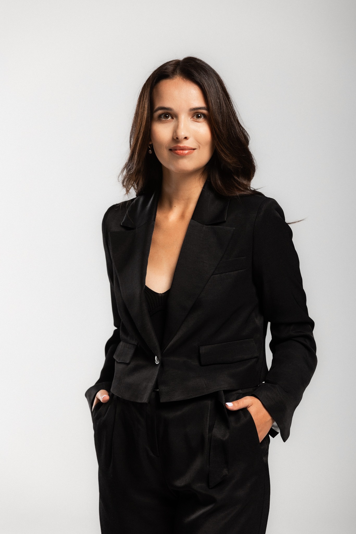 Portret kobiety w czarnym garniturze na jasnym tle. Uśmiech, dłonie w kieszeniach, profesjonalny wygląd. Idealna na sesję biznesową.