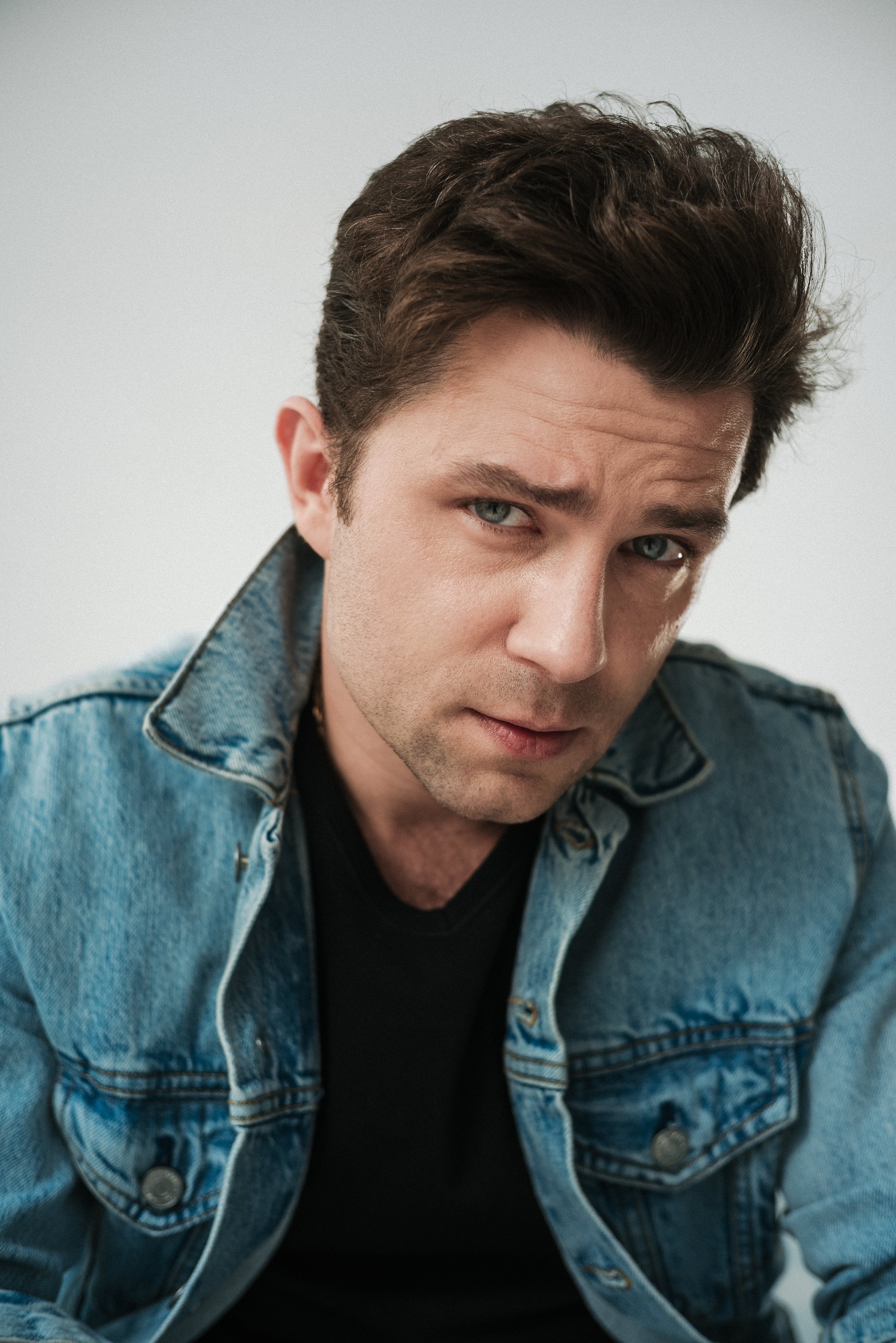 Portret mężczyzny z krótkimi, ciemnymi włosami, ubranego w dżinsową kurtkę i czarny t-shirt, na jasnym tle. Ujęcie z góry, podkreślające rysy twarzy i spojrzenie.