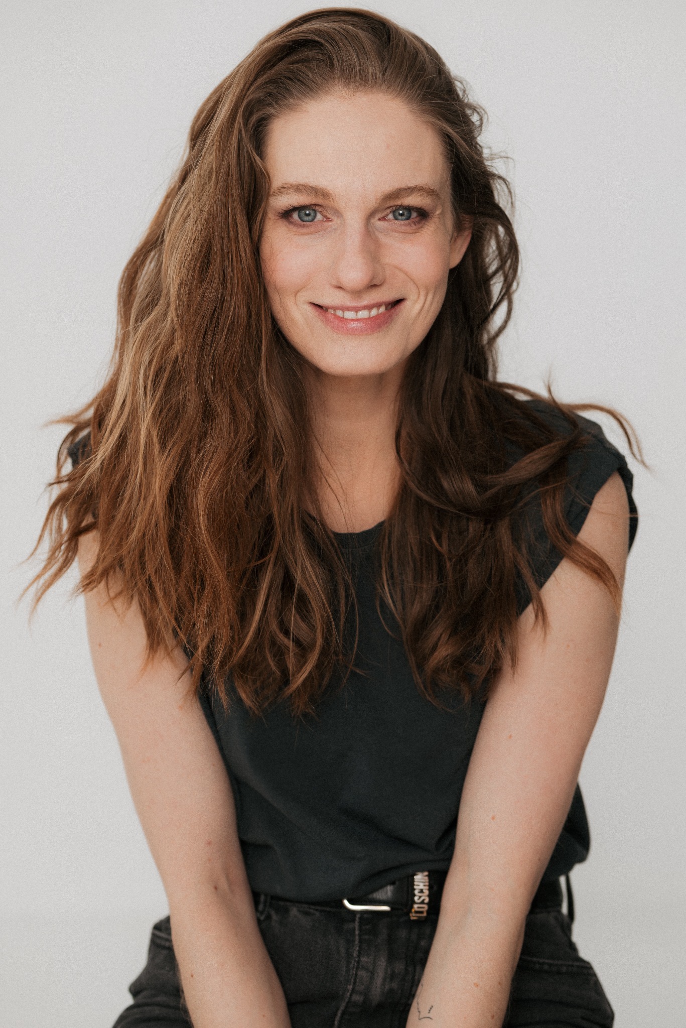 Portret uśmiechniętej kobiety o długich, brązowych włosach i niebieskich oczach, ubrana w ciemny t-shirt, na jasnym tle. Widoczny fragment paska z napisem.