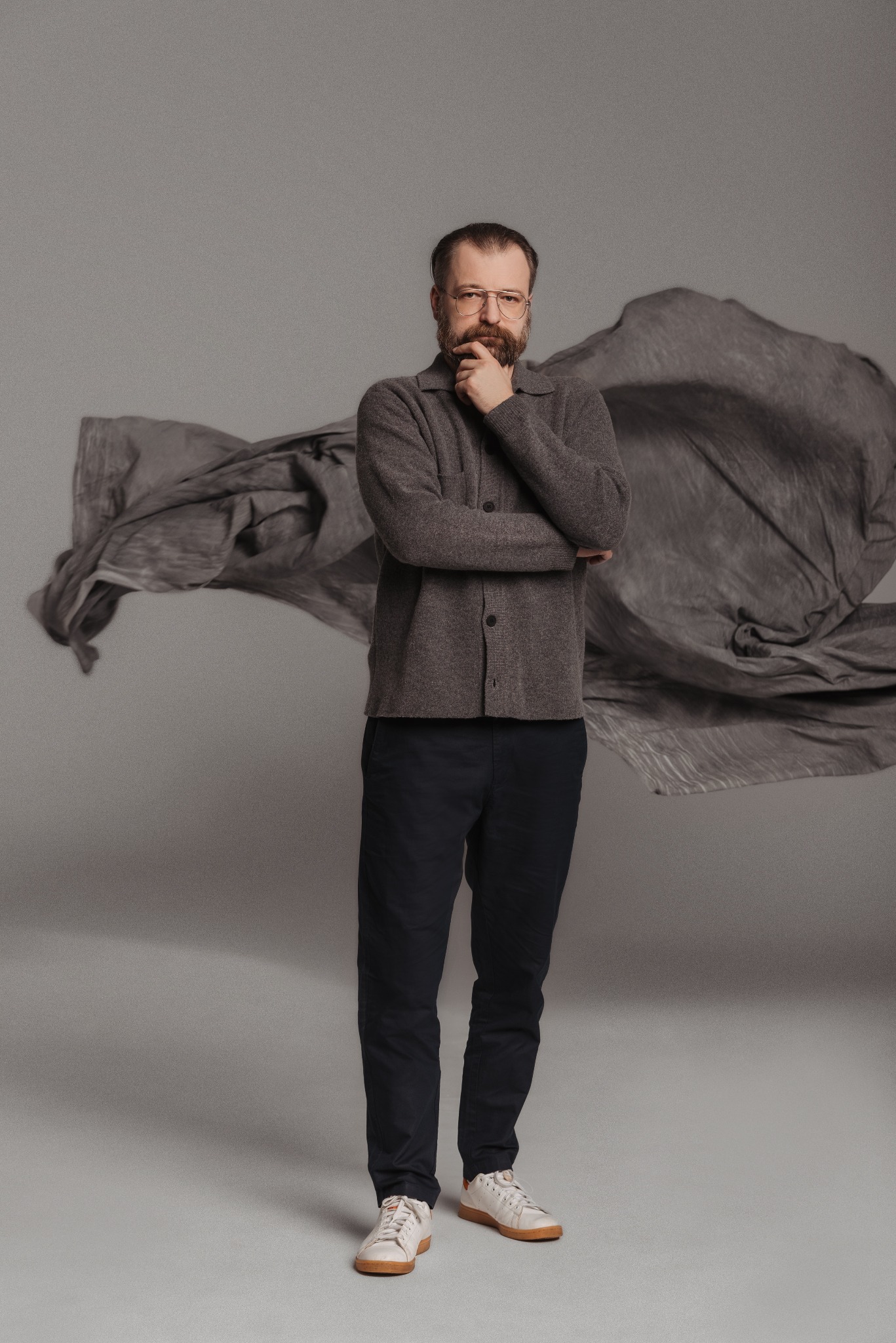Mężczyzna w okularach, z brodą, pozuje na szarym tle z unoszącą się tkaniną. Ubrany w sweter i spodnie, dłoń przy brodzie, patrzy w obiektyw. Sesja w studio.