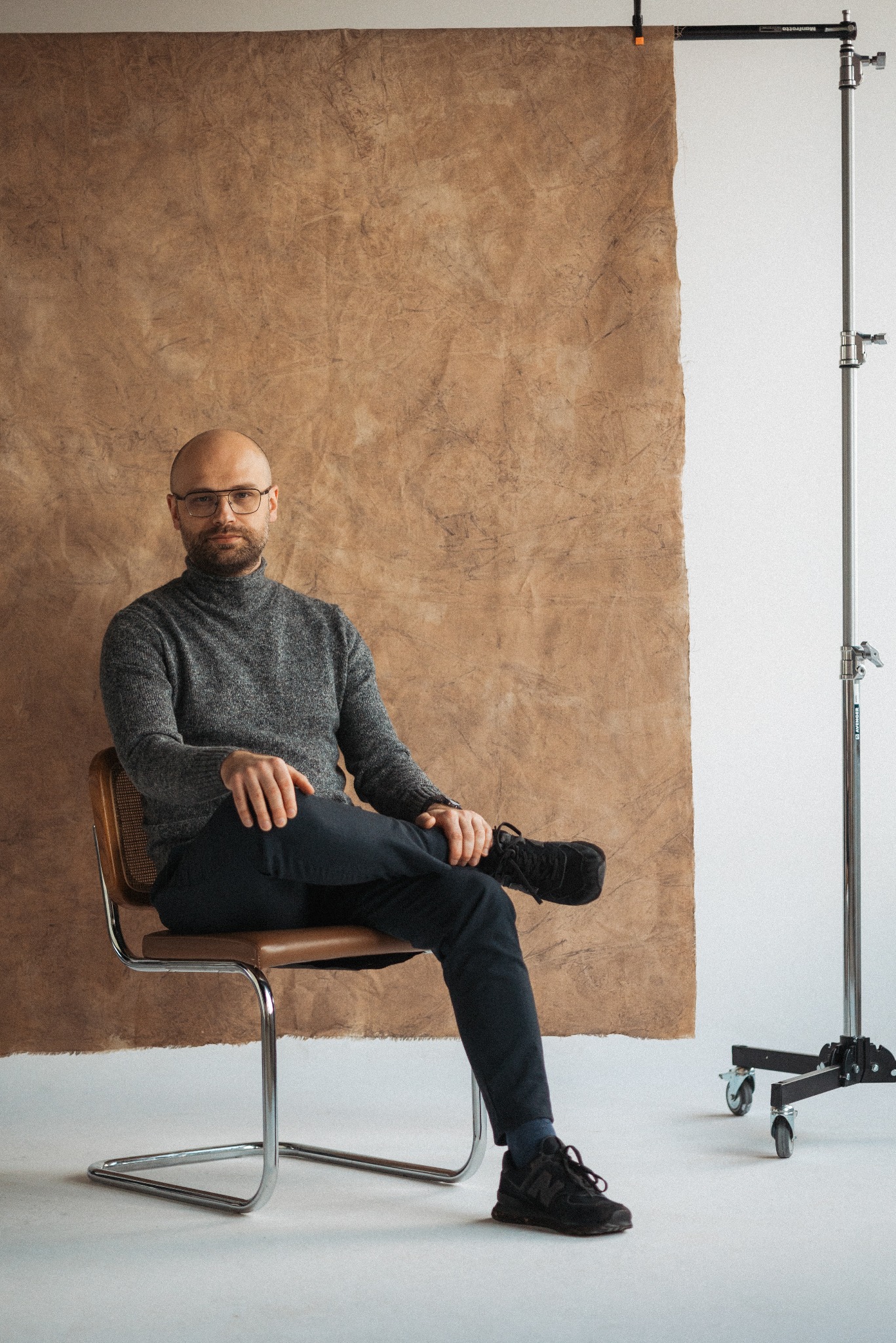 Mężczyzna w okularach i swetrze siedzi na krześle na tle studyjnym z brązowym tłem. Widoczny statyw oświetleniowy. Ujęcie portretowe w neutralnej tonacji.