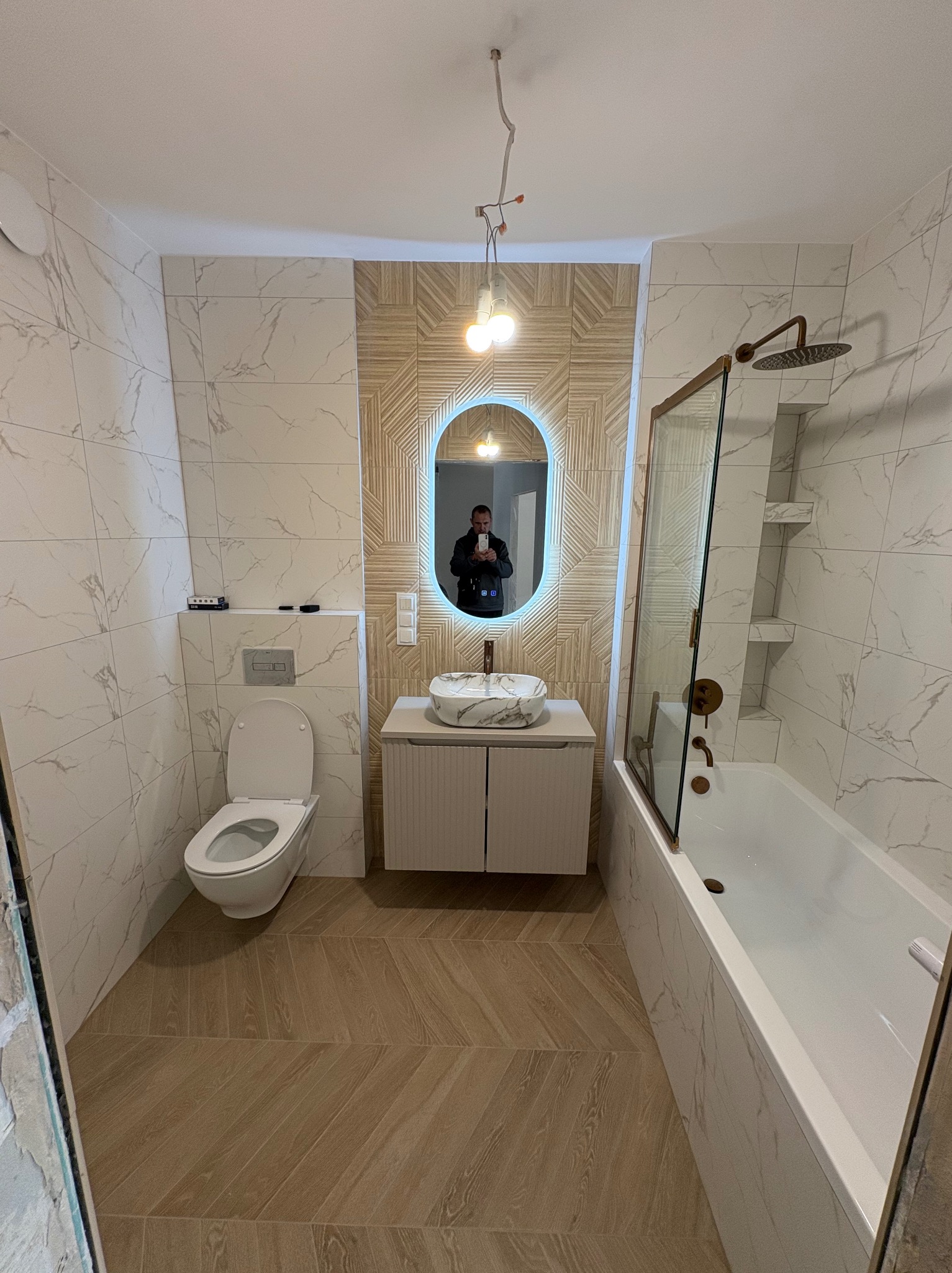 Wykończona łazienka we Wrocławiu z wanną, toaletą i umywalką na szafce. Białe płytki imitujące marmur na ścianach, drewnopodobne panele na podłodze, oświetlone lustro.