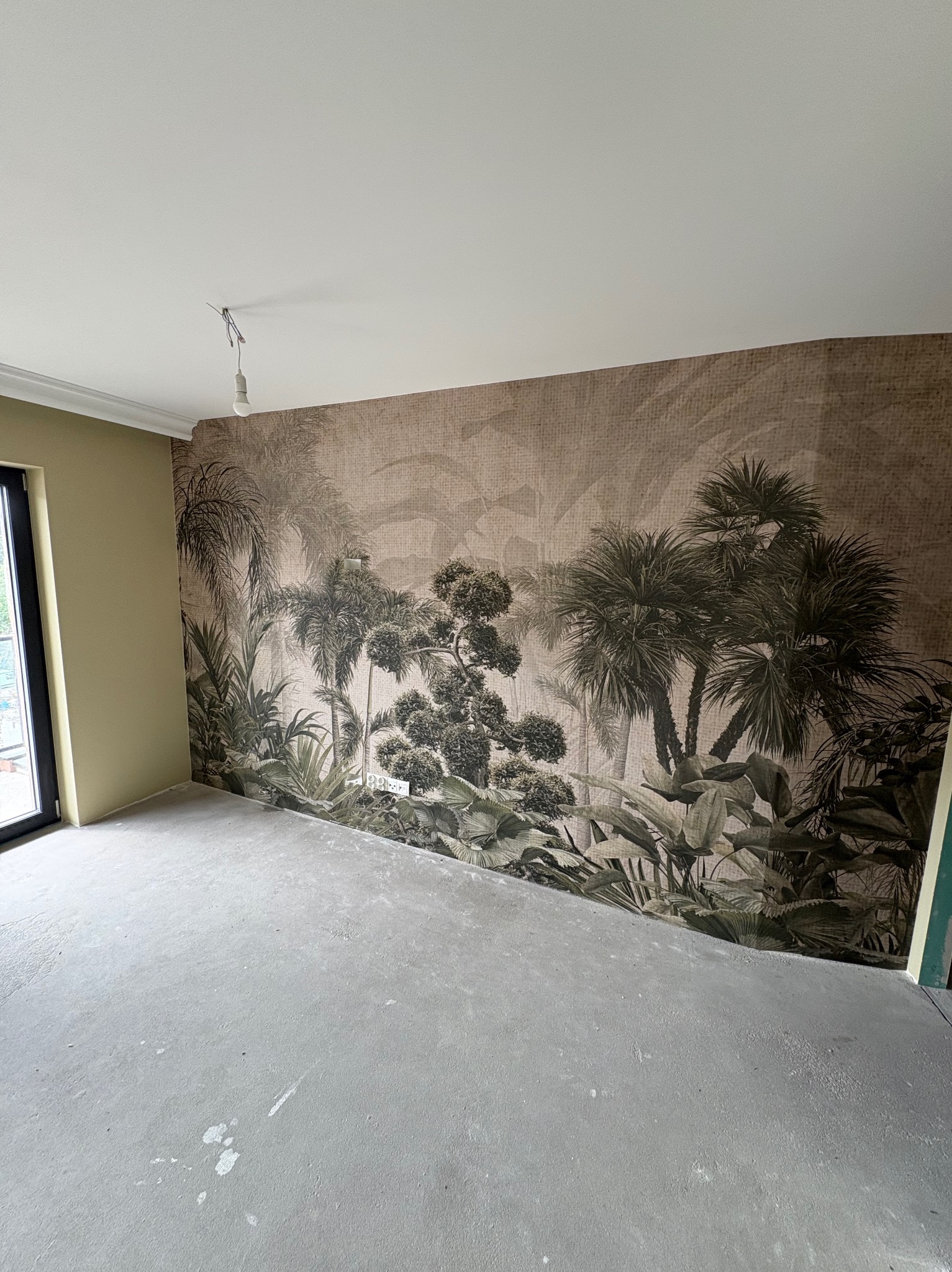 Pokój po tapetowaniu z motywem tropikalnym. Widoczna ściana z wzorem palm i roślin, surowa podłoga, okno i fragment pomalowanej ściany. Wystający kabel od oświetlenia.