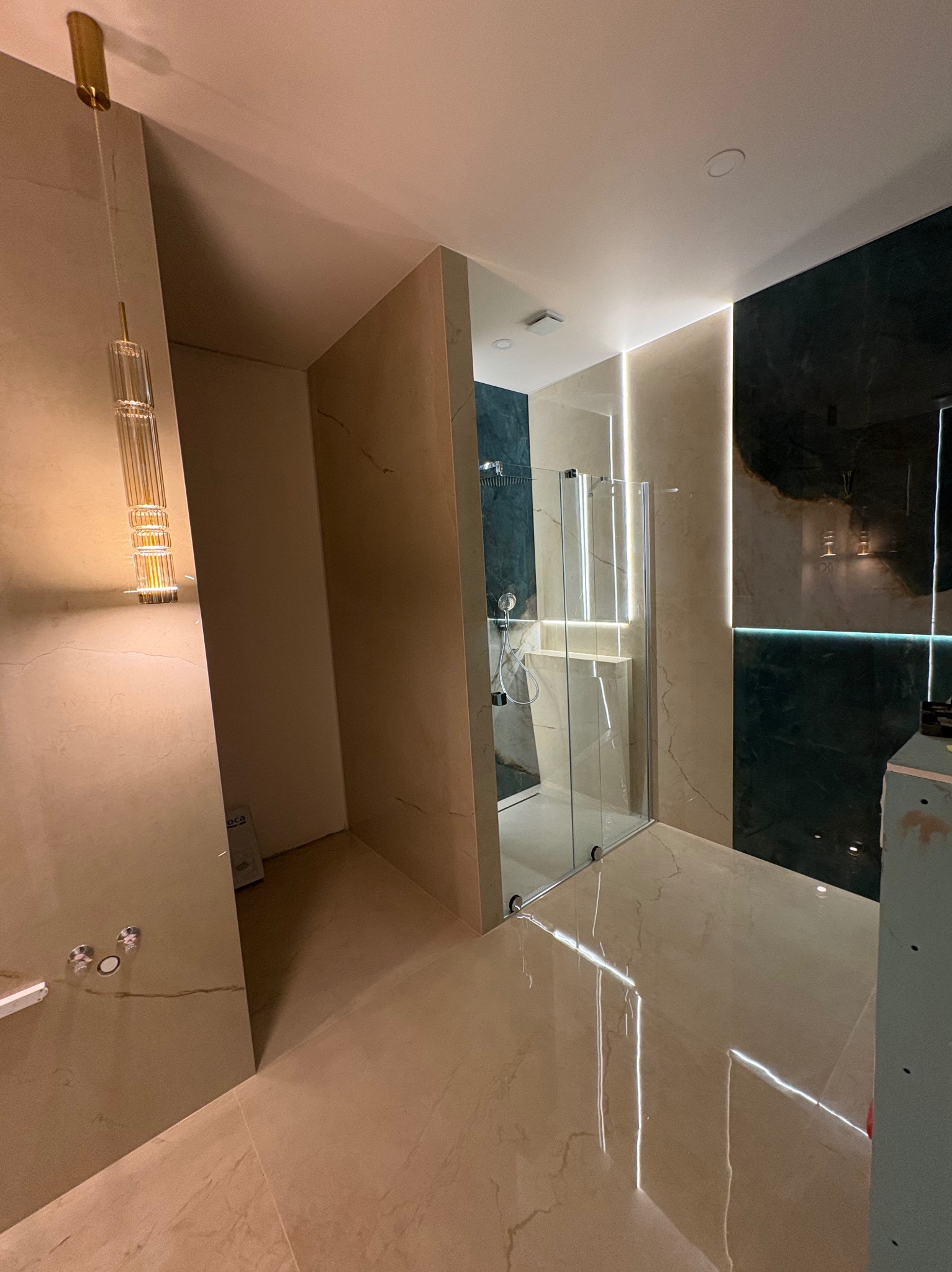 Nowoczesna łazienka z kabiną prysznicową typu walk-in, beżowe płytki imitujące marmur, akcenty ciemnej zieleni i oświetlenie LED. Widoczna designerska lampa wisząca.