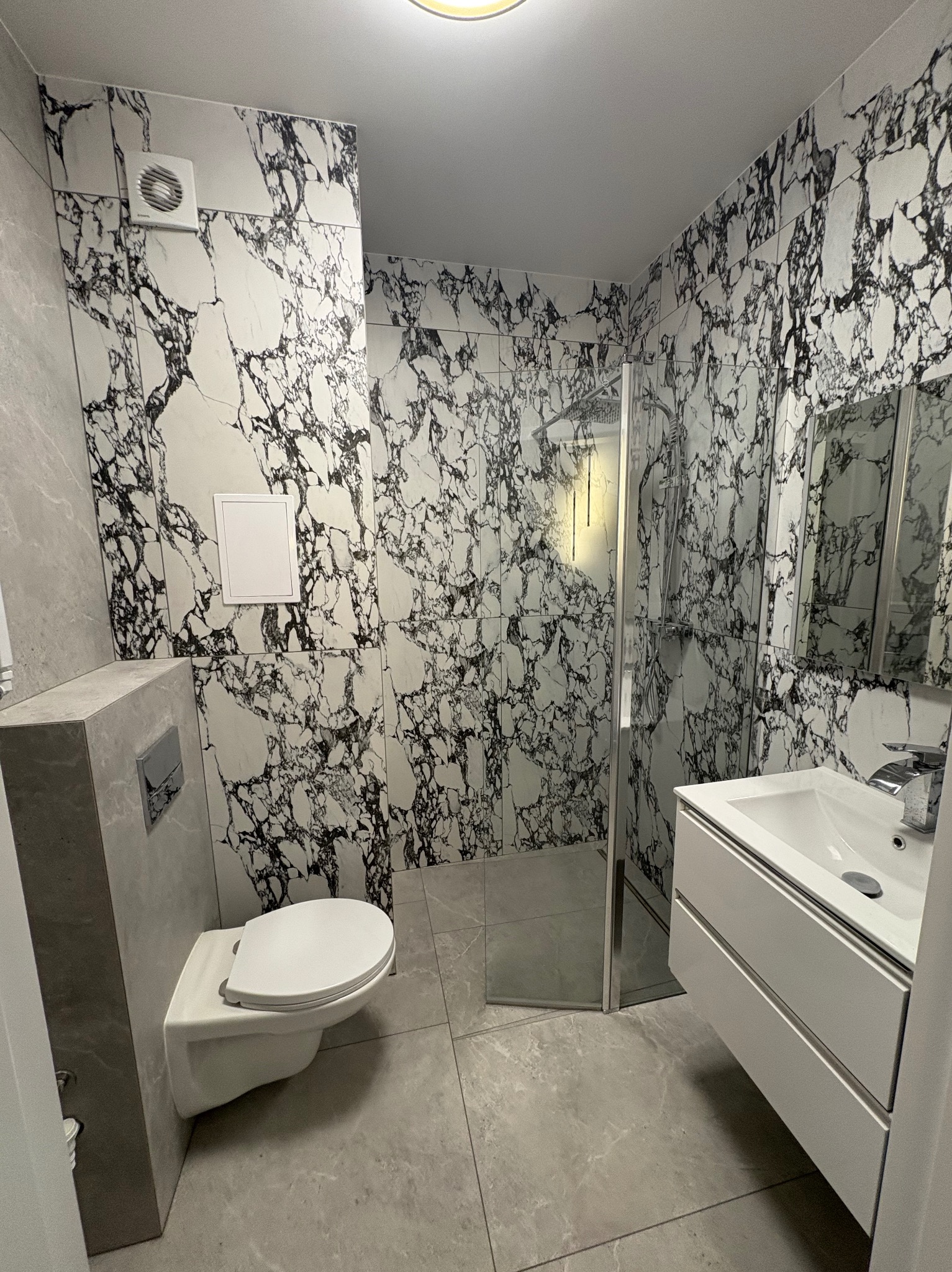 Nowoczesna łazienka z kabiną prysznicową, umywalką z szafką i podwieszaną toaletą. Ściany i podłoga wyłożone płytkami imitującymi marmur.