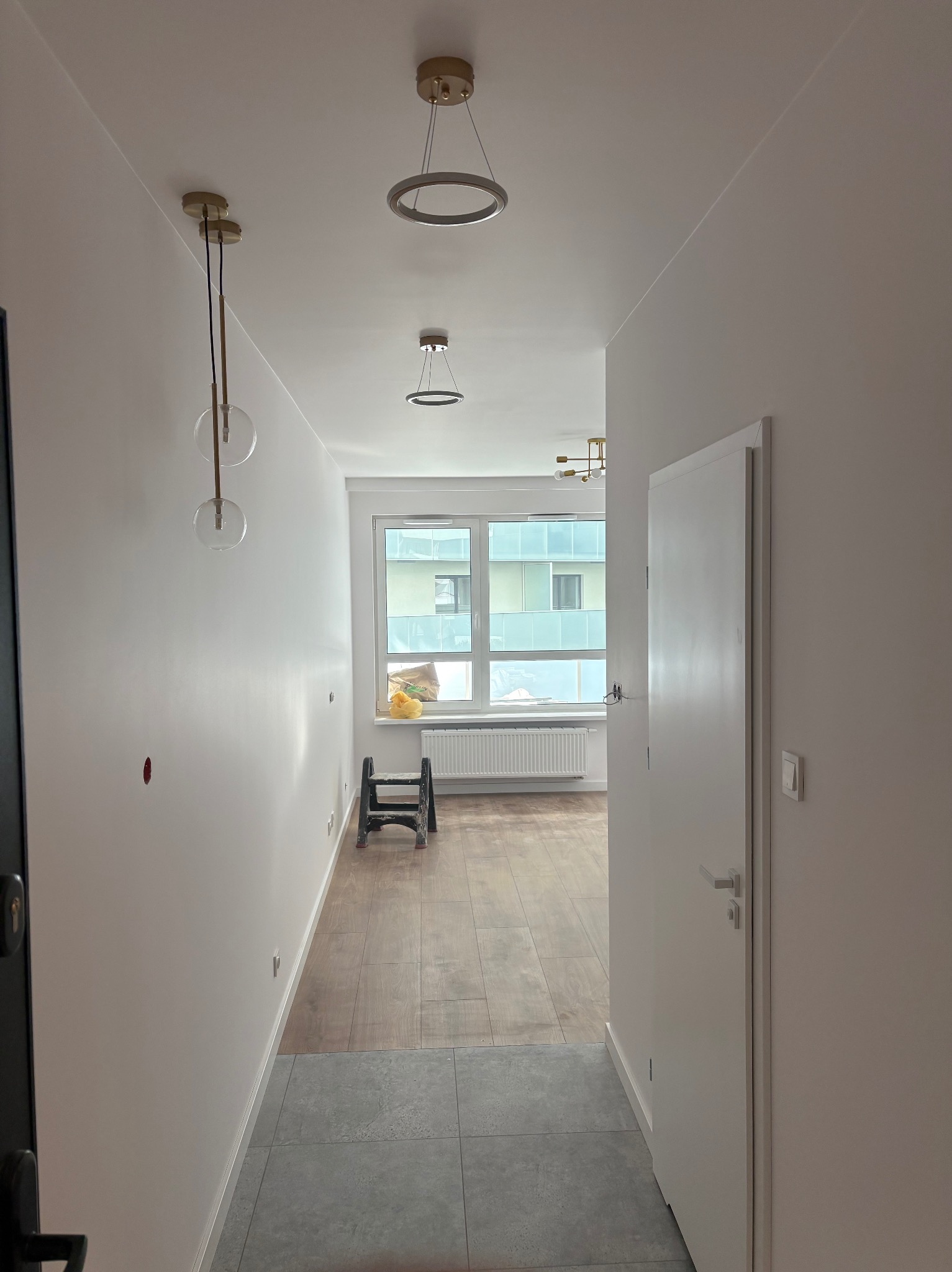 Nowoczesny korytarz z panelami podłogowymi przechodzącymi w szare płytki, białe ściany i sufit z trzema designerskimi lampami. Widoczne okno z widokiem na budynek.