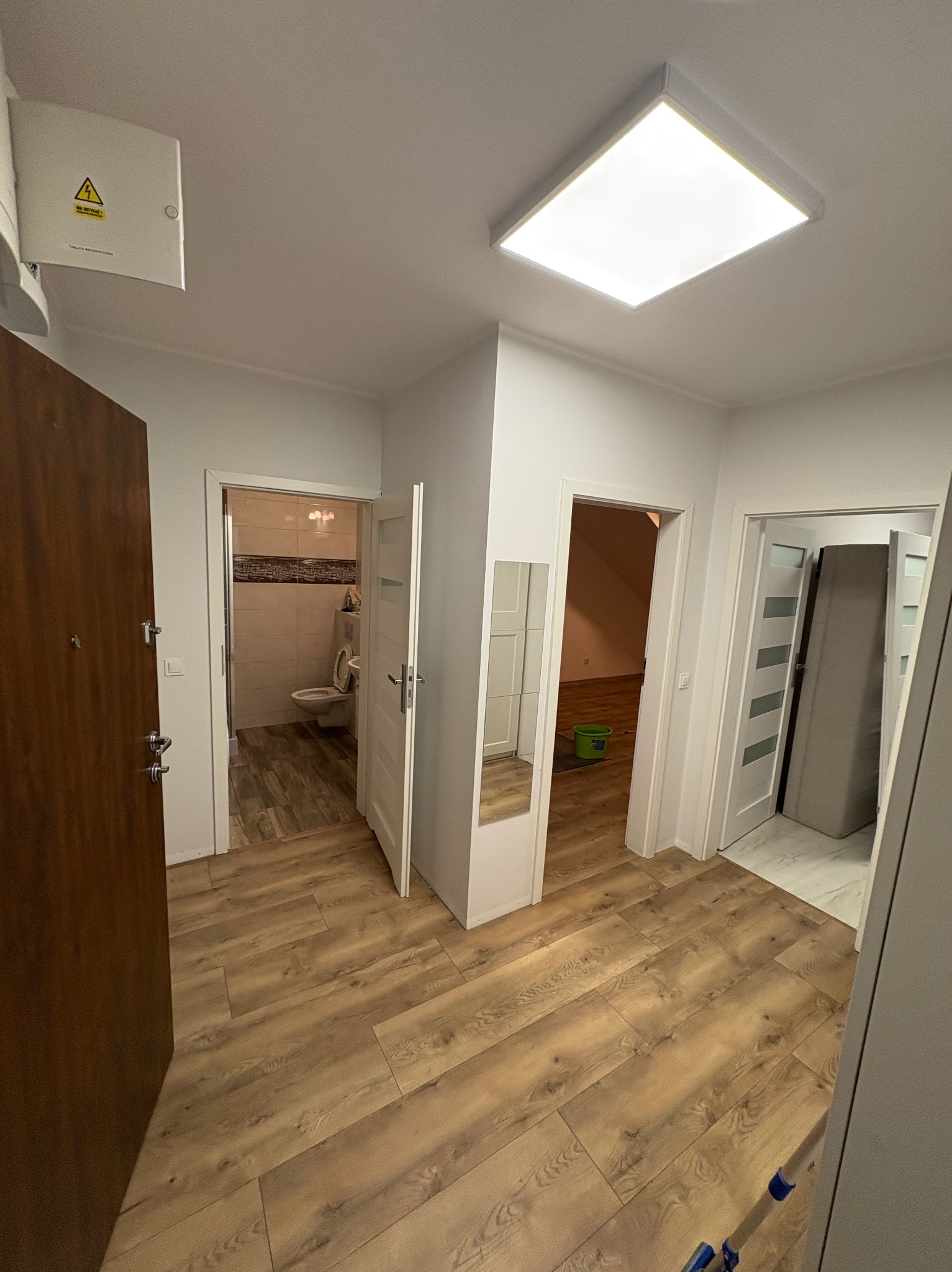 Jasne wnętrze mieszkania po remoncie: drewnopodobne panele podłogowe, białe ściany i drzwi, widok na łazienkę i inne pomieszczenia. Nowoczesne oświetlenie sufitowe.