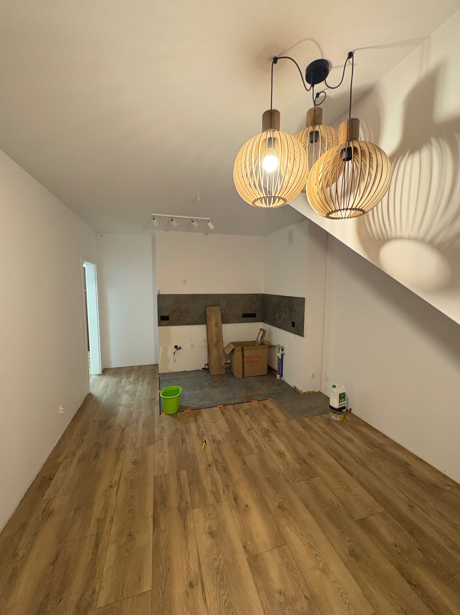 Jasne panele podłogowe ułożone w pokoju z trzema lampami wiszącymi. Częściowo wykończona ściana w tle. Widoczny fragment podłogi przed położeniem paneli.