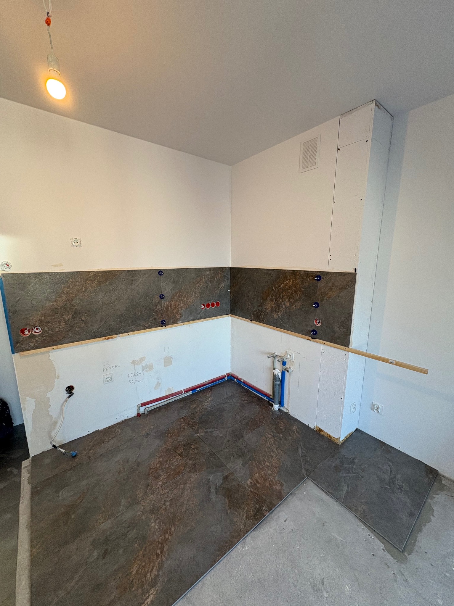 Fragment kuchni w trakcie prac. Częściowo ułożone płytki na podłodze i ścianie, widoczne instalacje wodne i elektryczne, przygotowane podłoże pod dalsze prace.