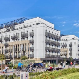 Bedroom Nieruchomości Świnoujście i Szczecin - Nowoczesny, biały budynek apartamentowy z czarnymi balkonami i spiralnymi schodami na dachu, widziany z poziomu ulicy z przechodniami i rowerami. Słoneczny dzień.