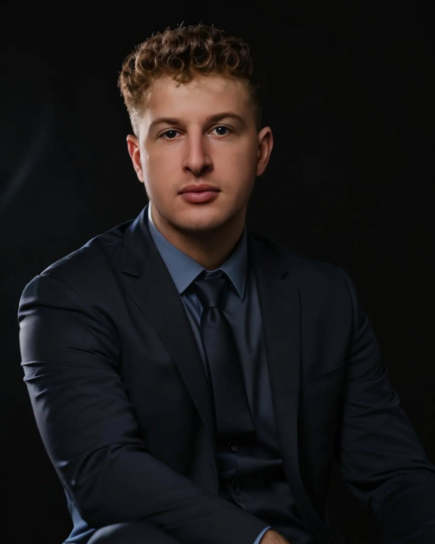 Portret młodego mężczyzny w ciemnym garniturze i niebieskiej koszuli na czarnym tle, z delikatnym uśmiechem.