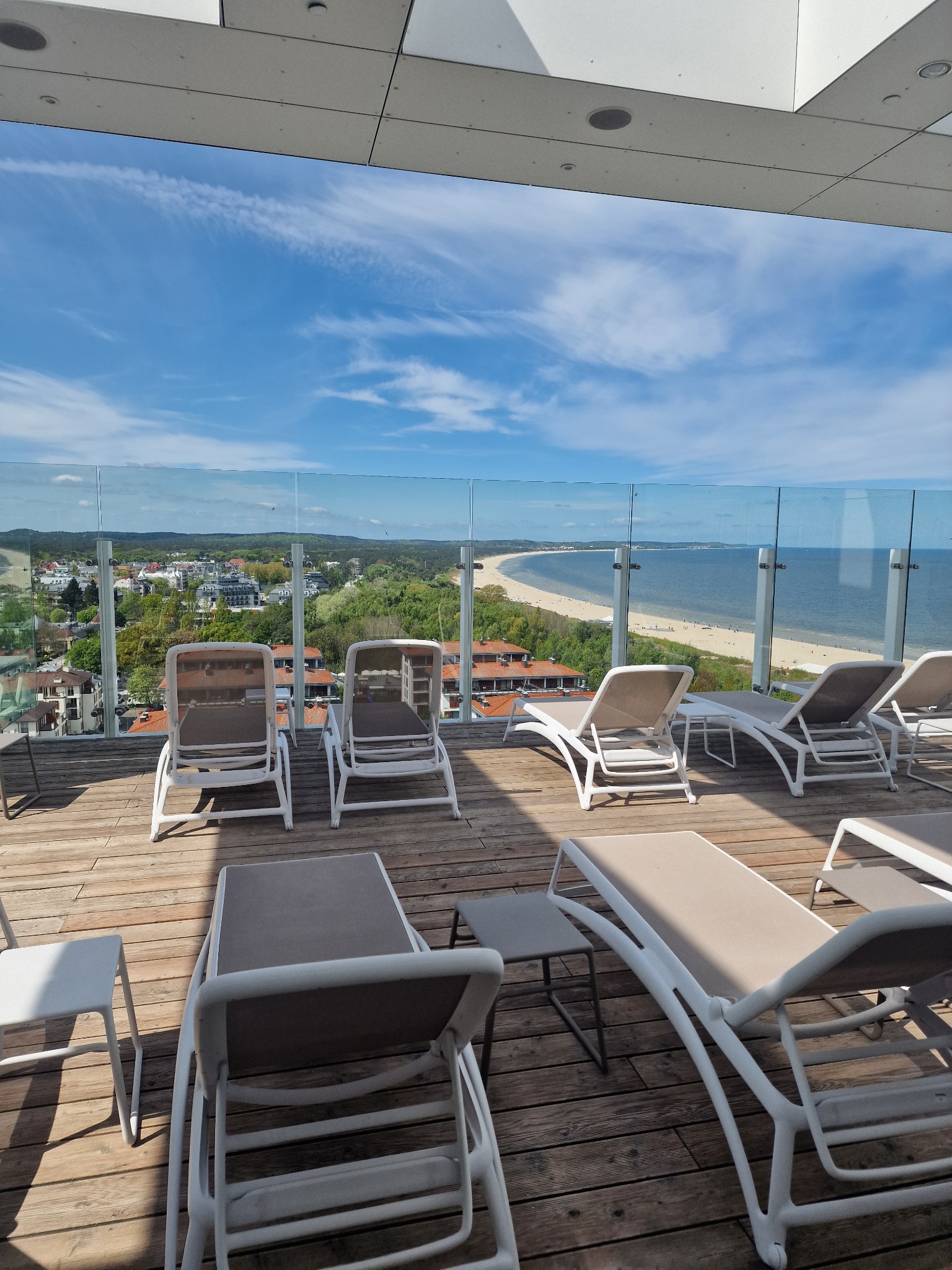 Drewniany taras z leżakami i widokiem na morze z wysokiego piętra. W tle budynki i plaża. Błękitne niebo z chmurami. Przeszklenia zapewniają ochronę.