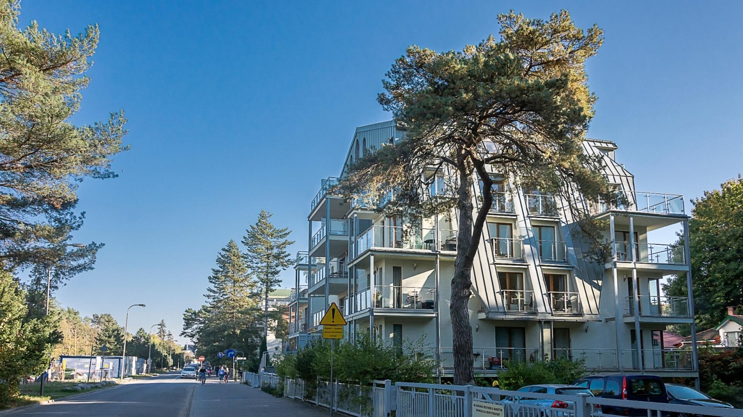 Nowoczesny apartamentowiec z balkonami w otoczeniu zieleni miejskiej, widok z poziomu ulicy. Uwagę zwraca charakterystyczny kształt dachu i szklane balustrady.
