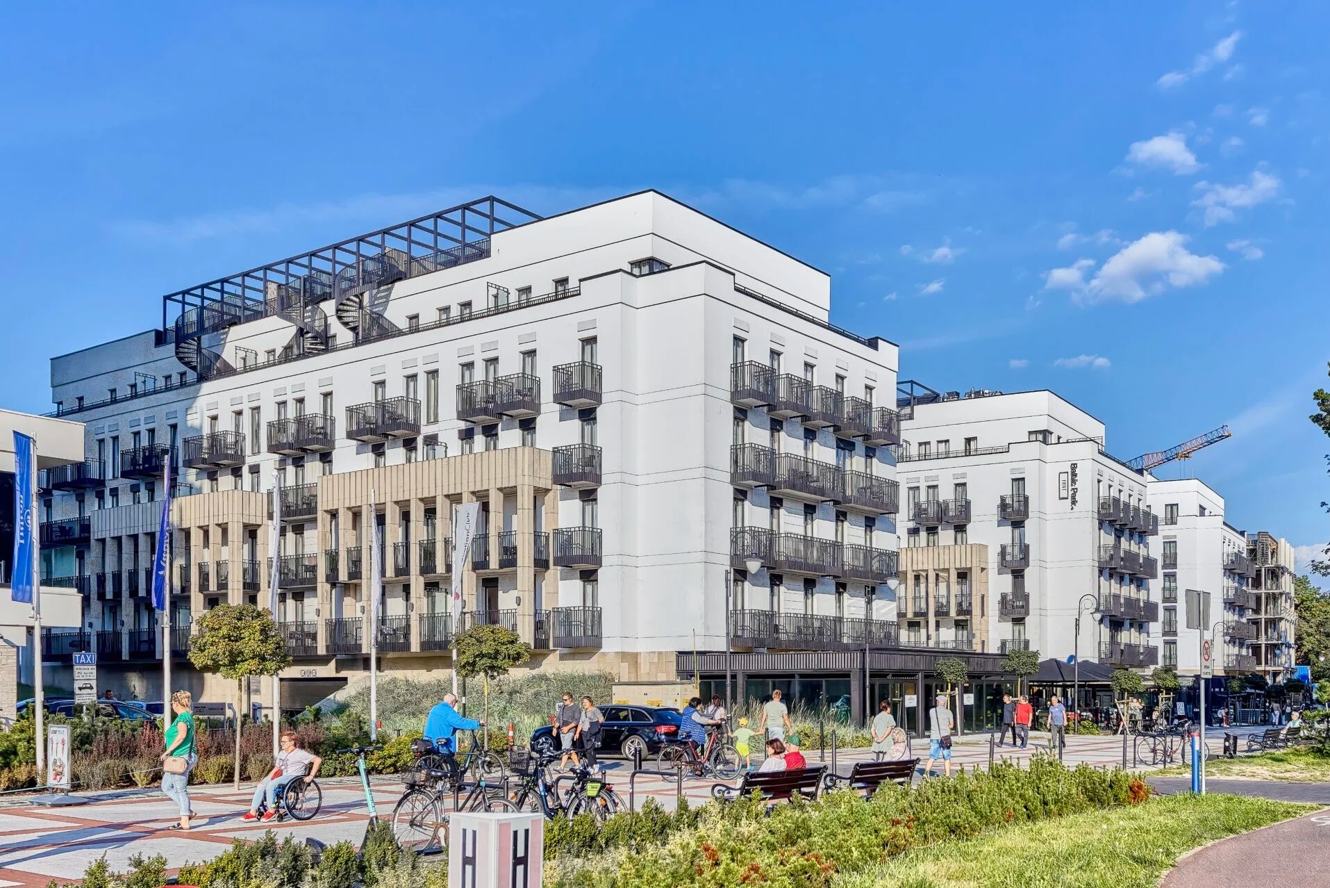 Nowoczesny, biały budynek apartamentowy z czarnymi balkonami i spiralnymi schodami na dachu, widziany z poziomu ulicy z przechodniami i rowerami. Słoneczny dzień.