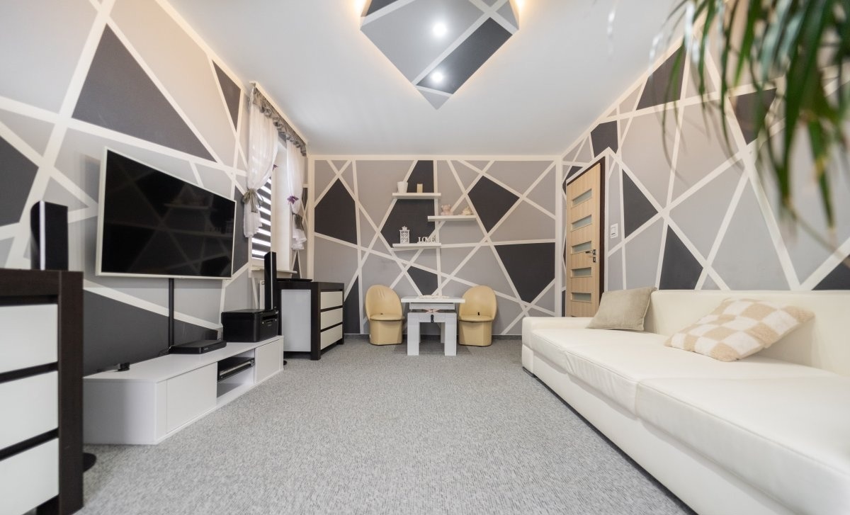 Nowoczesny salon z geometrycznym wzorem na ścianach w odcieniach szarości i bieli, jasna sofa i meble, okno z roletami, roślina w rogu.