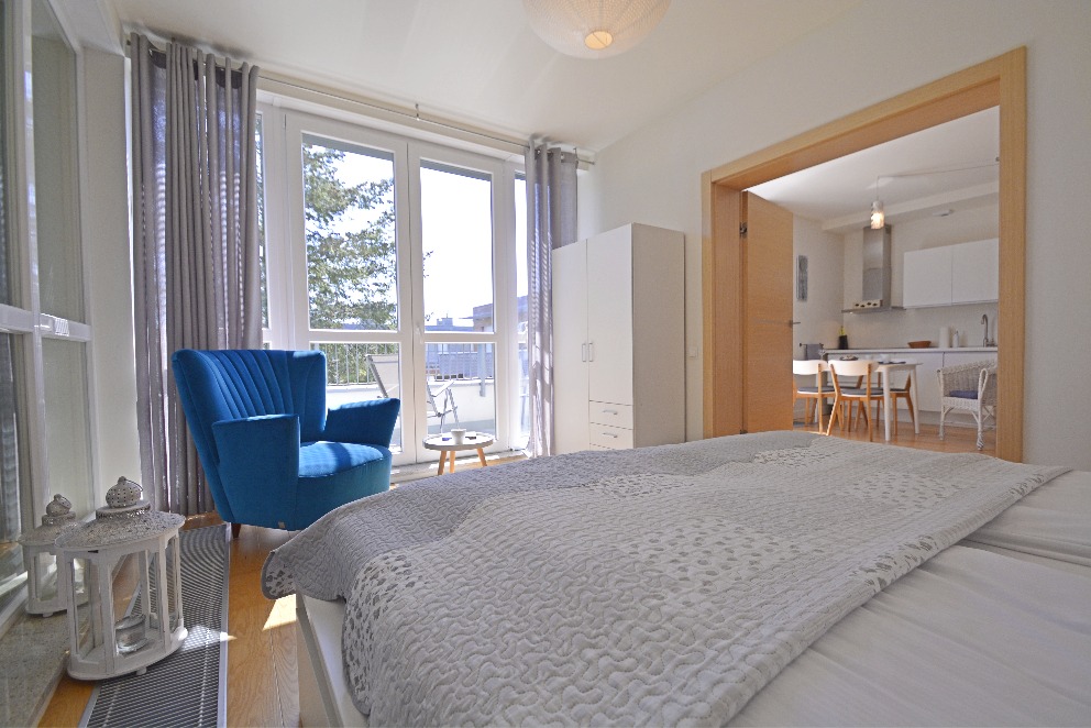 Jasne wnętrze: błękitny fotel w sypialni z balkonem i widokiem na miasto, przez otwarte drzwi widać kuchnię. Nowoczesny design, neutralna kolorystyka, naturalne światło.