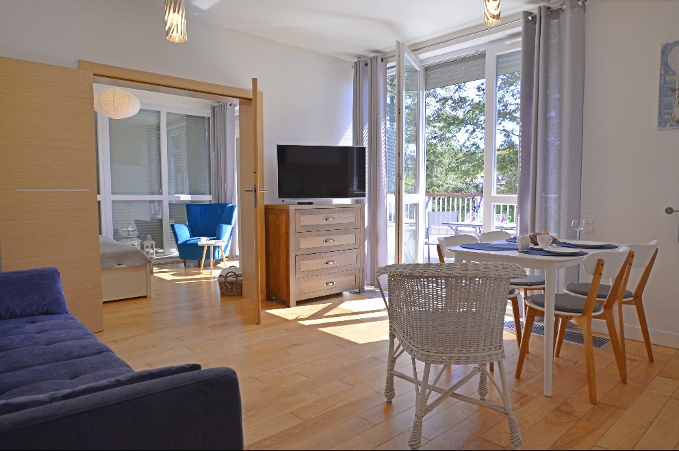 Jasne wnętrze apartamentu z drewnianą podłogą, niebieską sofą i fotelem, komodą z telewizorem oraz stołem z krzesłami, balkon z widokiem na zieleń.