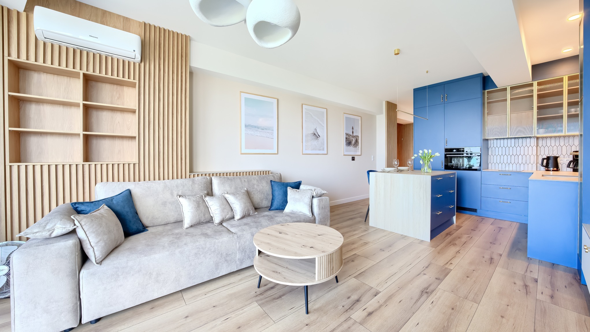 Jasne wnętrze mieszkania z drewnianą ścianą, szarą sofą z poduszkami, okrągłym stolikiem i niebieską zabudową kuchenną. Na ścianie wiszą obrazy z motywami plażowymi.