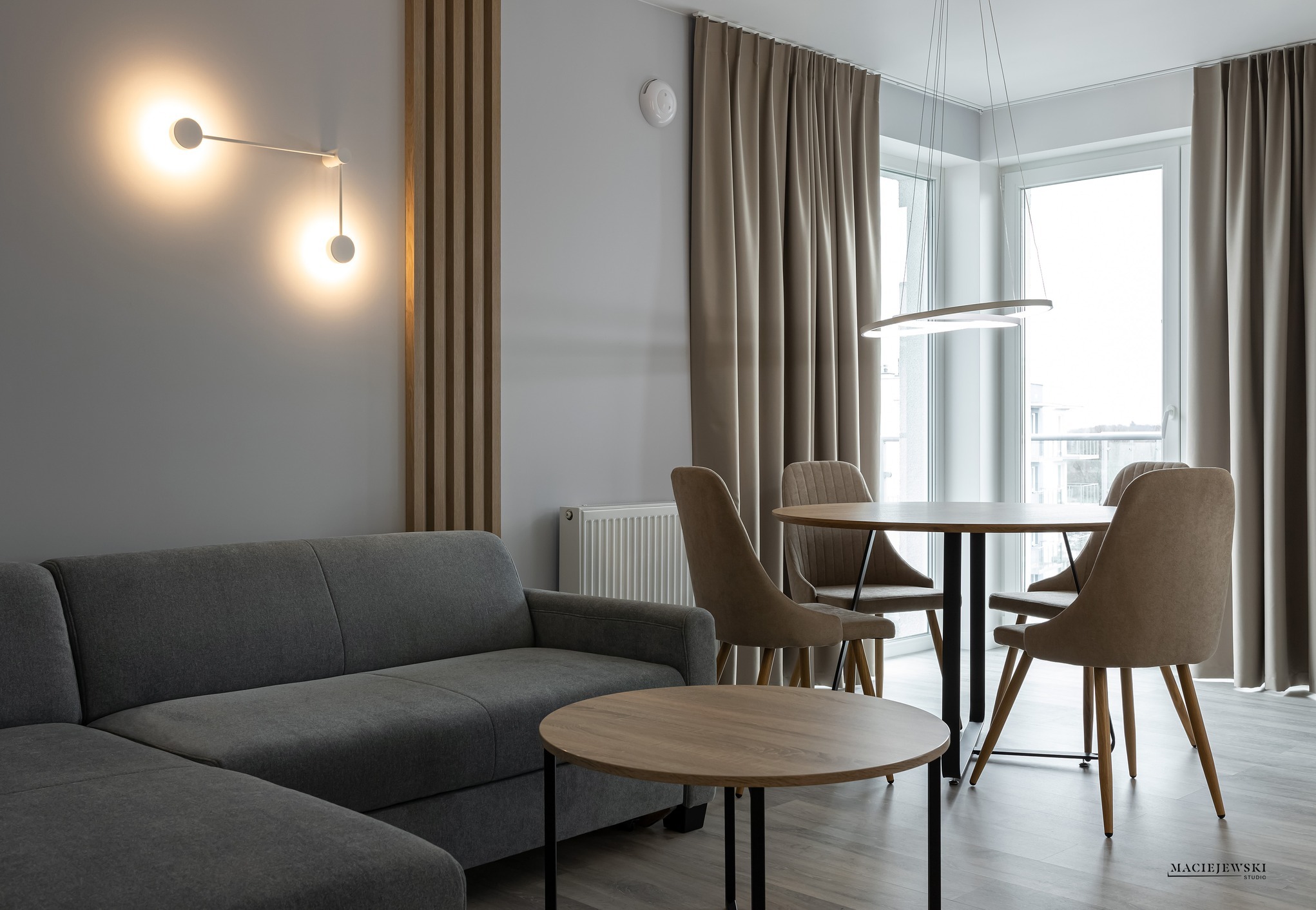 Nowoczesne wnętrze salonu w Świnoujściu: szara sofa, okrągły stół z krzesłami, drewniane lamele na ścianie i designerskie oświetlenie, tworzące przytulną atmosferę.