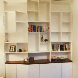 BRUS KRZYSZTOF GREEN MIND - Biała, narożna szafa wnękowa z otwartymi półkami i ciemnym blatem, wypełniona książkami i dekoracjami. Minimalistyczny design, dopasowana do kąta pomieszczenia.