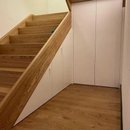 BRUS KRZYSZTOF GREEN MIND - Drewniane schody z zabudową wnękową pod nimi. Białe fronty szafek wkomponowane w przestrzeń pod schodami, drewniana podłoga. Funkcjonalne wykorzystanie przestrzeni.