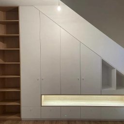 BRUS KRZYSZTOF GREEN MIND - Biała zabudowa wnęki pod skosem z szafkami, szufladami i półkami, oświetlona listwą LED, z drewnianym regałem z lewej strony. Zabudowa dopasowana do skosu dachu.