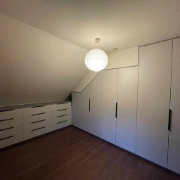 BRUS KRZYSZTOF GREEN MIND - Biała szafa wnękowa z szufladami pod skosem, czarne uchwyty, drewniana podłoga, oświetlenie punktowe. Minimalistyczny design w jasnym wnętrzu.