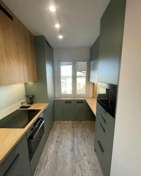 Wąska kuchnia w odcieniach zieleni i drewna, z oknem na końcu. Uwagę zwraca nietypowy układ szafek dolnych pod oknem i drewniana podłoga imitująca panele.