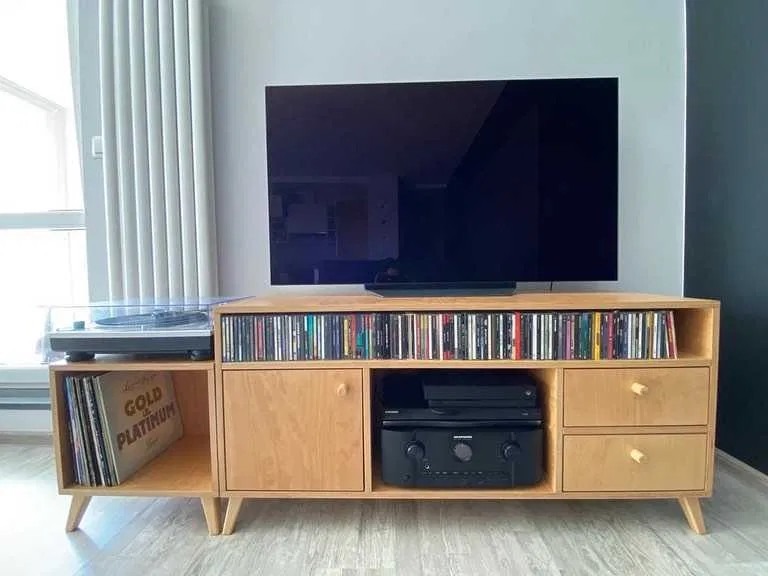 Jasny, drewniany regał RTV na nóżkach, wypełniony płytami CD i winylami, z gramofonem i sprzętem audio. Nad nim telewizor. Minimalistyczny design.