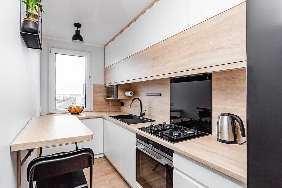 Funkcjonalna kuchnia z białymi szafkami i drewnianymi blatami, czarną armaturą i składanym stołem przy oknie. Minimalistyczny design z rośliną na półce.