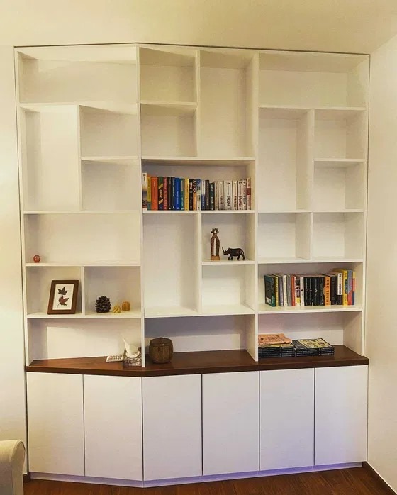 Biała, narożna szafa wnękowa z otwartymi półkami i ciemnym blatem, wypełniona książkami i dekoracjami. Minimalistyczny design, dopasowana do kąta pomieszczenia.