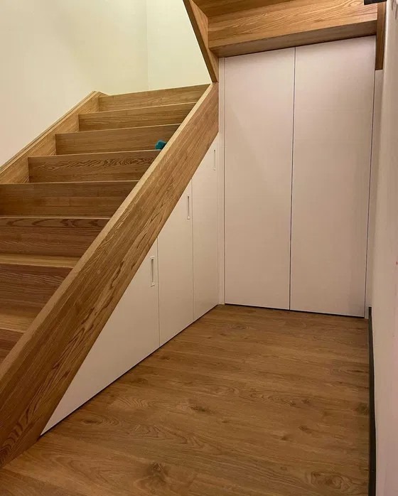 Drewniane schody z zabudową wnękową pod nimi. Białe fronty szafek wkomponowane w przestrzeń pod schodami, drewniana podłoga. Funkcjonalne wykorzystanie przestrzeni.