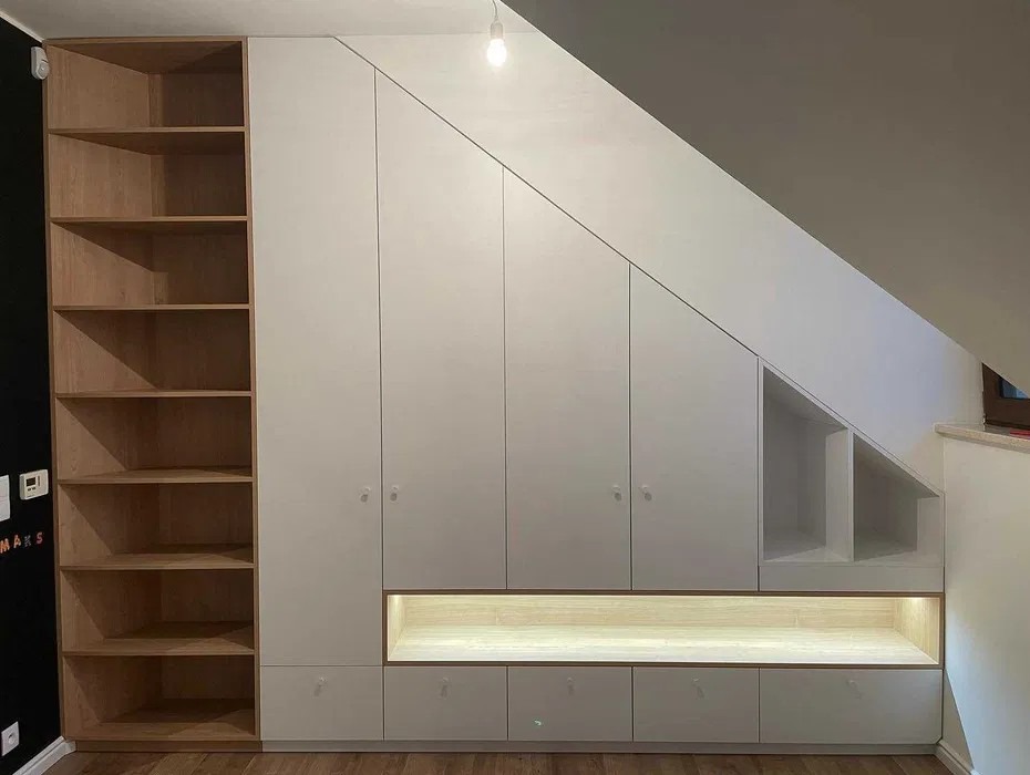Biała zabudowa wnęki pod skosem z szafkami, szufladami i półkami, oświetlona listwą LED, z drewnianym regałem z lewej strony. Zabudowa dopasowana do skosu dachu.
