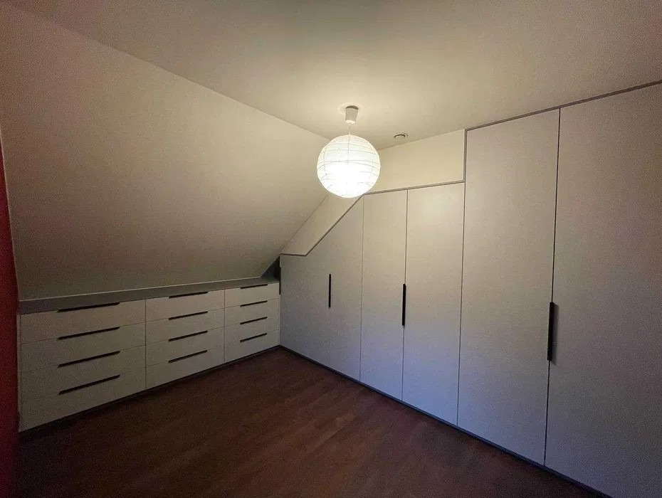 Biała szafa wnękowa z szufladami pod skosem, czarne uchwyty, drewniana podłoga, oświetlenie punktowe. Minimalistyczny design w jasnym wnętrzu.