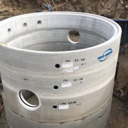 HYDRA - Betonowy, prefabrykowany krąg szamba z widocznymi otworami i oznaczeniami producenta Ecol-Unicon, ustawiony w wykopie, z ubijarką w tle na brzegu wykopu.