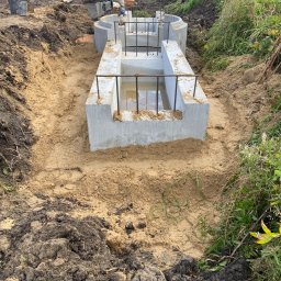 HYDRA - Montaż betonowego szamba w wykopie, widoczny pracownik z zagęszczarką, ujęcie z góry na częściowo wypełniony wodą zbiornik z wystającymi prętami zbrojeniowymi.