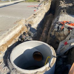 HYDRA - Betonowy krąg studni kanalizacyjnej z widoczną rurą wewnątrz, wykop z rurą w ziemi, ubijarka wibracyjna i fragment koparki w tle.