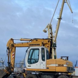 HYDRA - Żółto-biała koparka Liebherr 314 z dodatkowym ramieniem dźwigu na tle pochmurnego nieba i portowych konstrukcji.