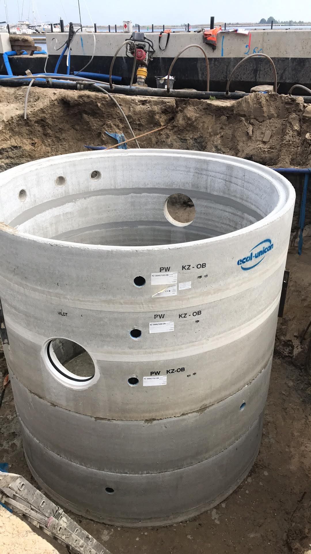 Betonowy, prefabrykowany krąg szamba z widocznymi otworami i oznaczeniami producenta Ecol-Unicon, ustawiony w wykopie, z ubijarką w tle na brzegu wykopu.