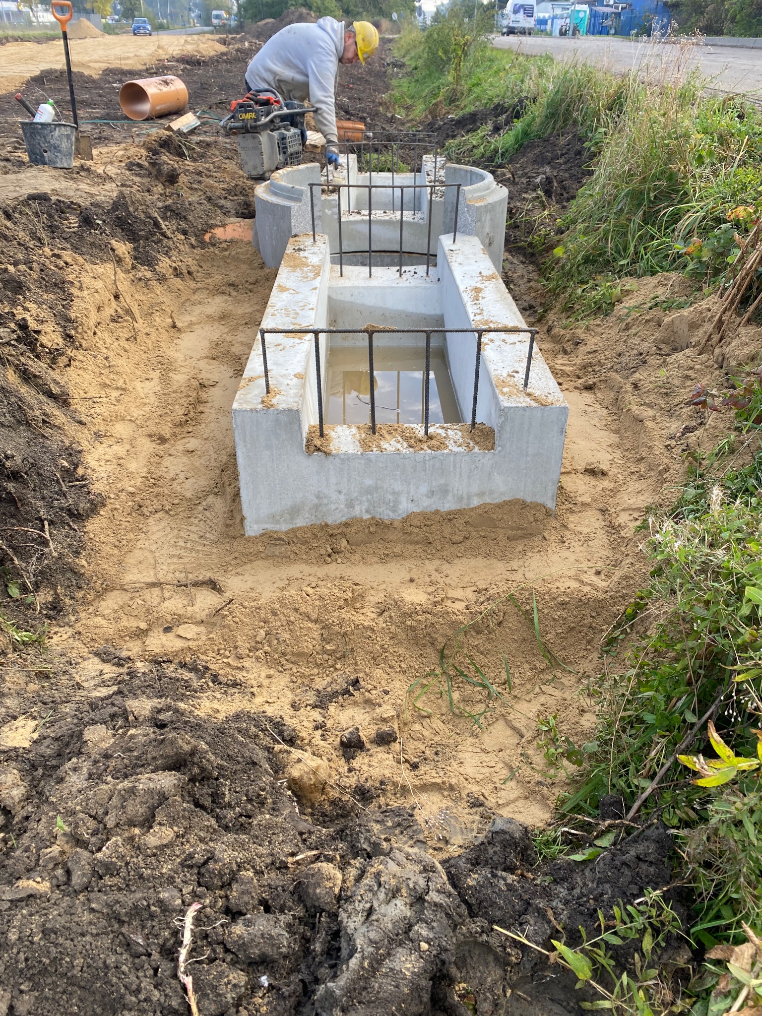 Montaż betonowego szamba w wykopie, widoczny pracownik z zagęszczarką, ujęcie z góry na częściowo wypełniony wodą zbiornik z wystającymi prętami zbrojeniowymi.