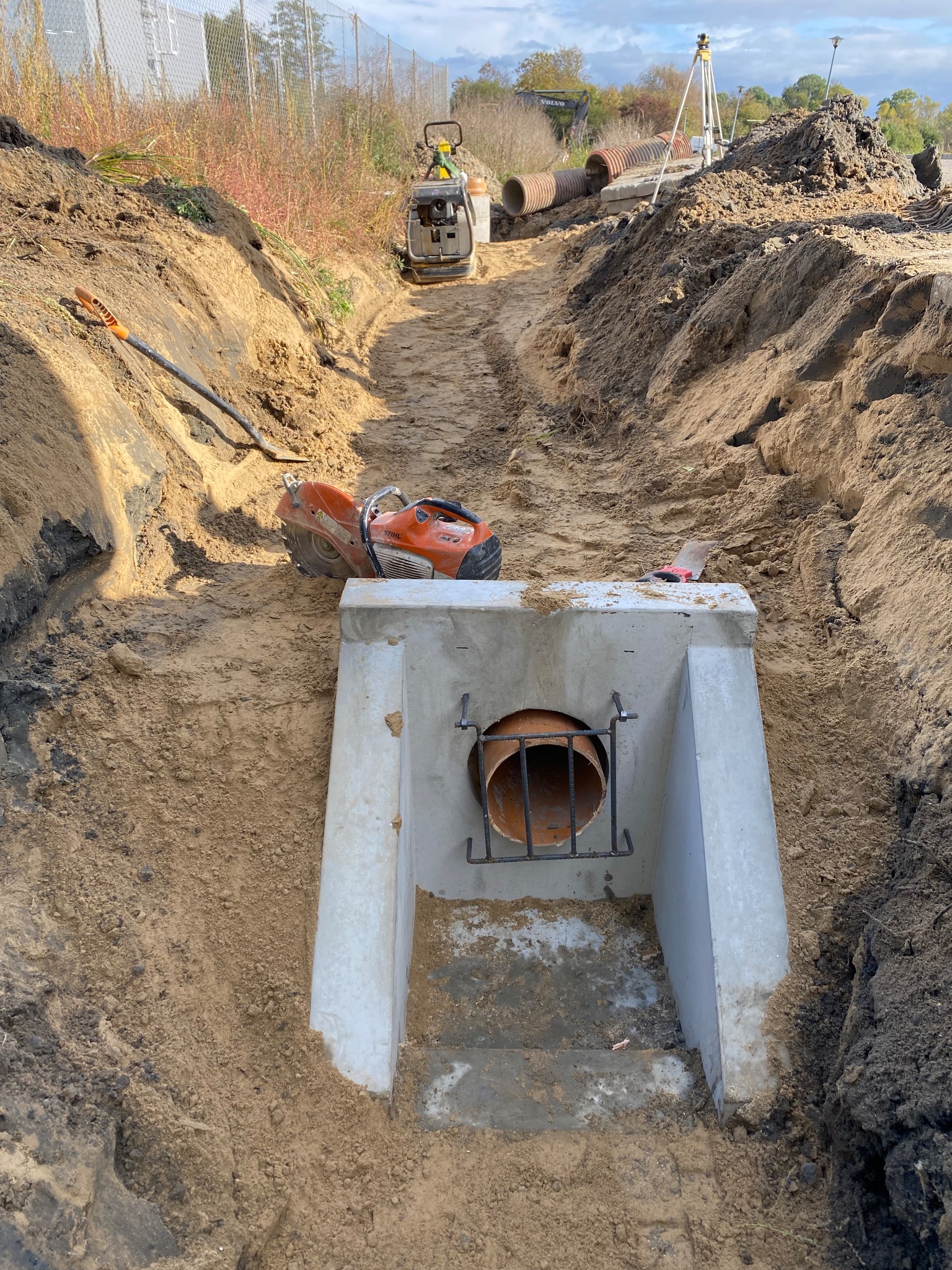 Betonowa studzienka z rurą w wykopie, pomarańczowa piła do betonu, ubijarka i ułożone rury w tle, widok z góry na plac budowy.