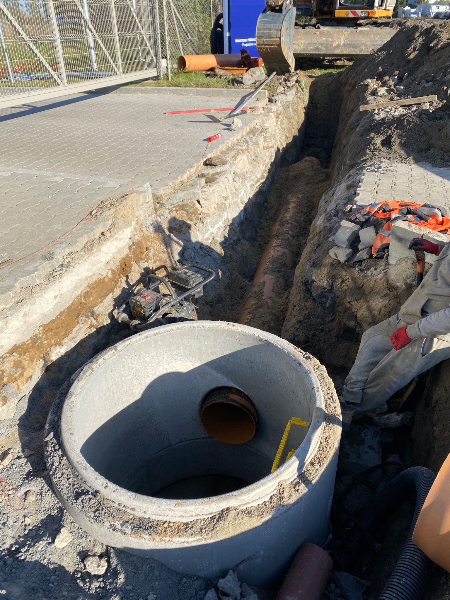 Betonowy krąg studni kanalizacyjnej z widoczną rurą wewnątrz, wykop z rurą w ziemi, ubijarka wibracyjna i fragment koparki w tle.