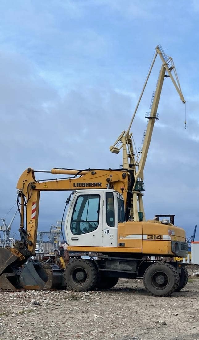Żółto-biała koparka Liebherr 314 z dodatkowym ramieniem dźwigu na tle pochmurnego nieba i portowych konstrukcji.